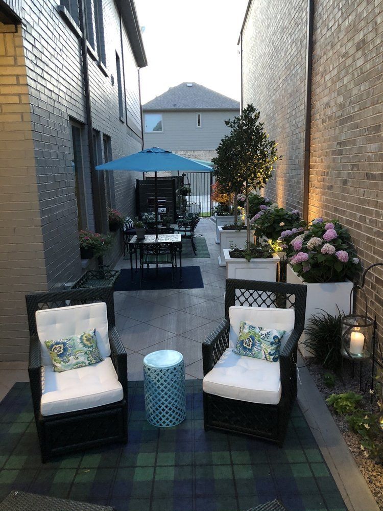 Outdoor patio with two black chairs, teal table, umbrella, and potted flowers between brick walls.