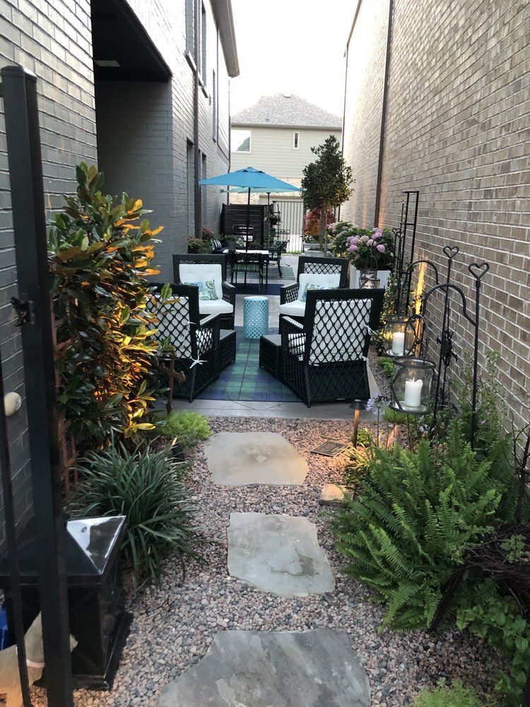 Narrow outdoor patio with black wicker chairs, potted plants, string lights, and a stone path between brick walls