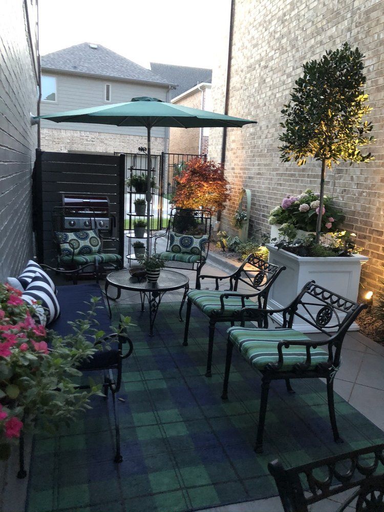 Small patio with black metal furniture, green umbrella, potted flowers, and plaid outdoor rug between buildings