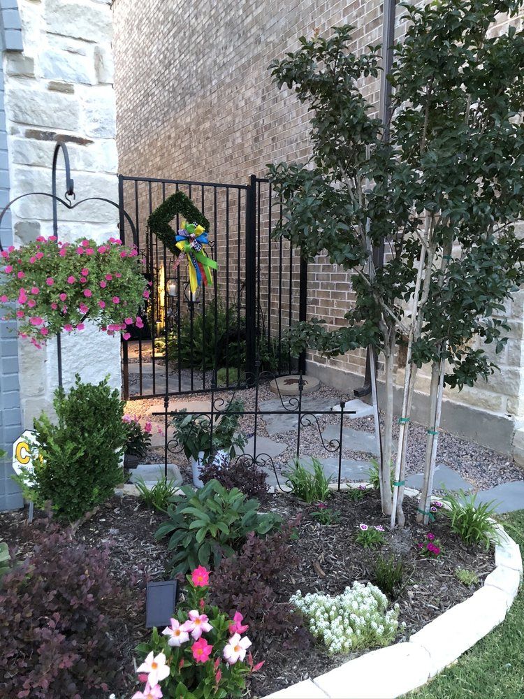 Flower bed with pink and white blooms, a hanging basket, and a black metal fence by a stone wall
