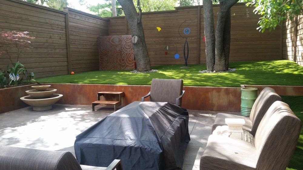 Backyard patio with covered furniture, fire pit, brick walls, and a grassy raised garden area with trees