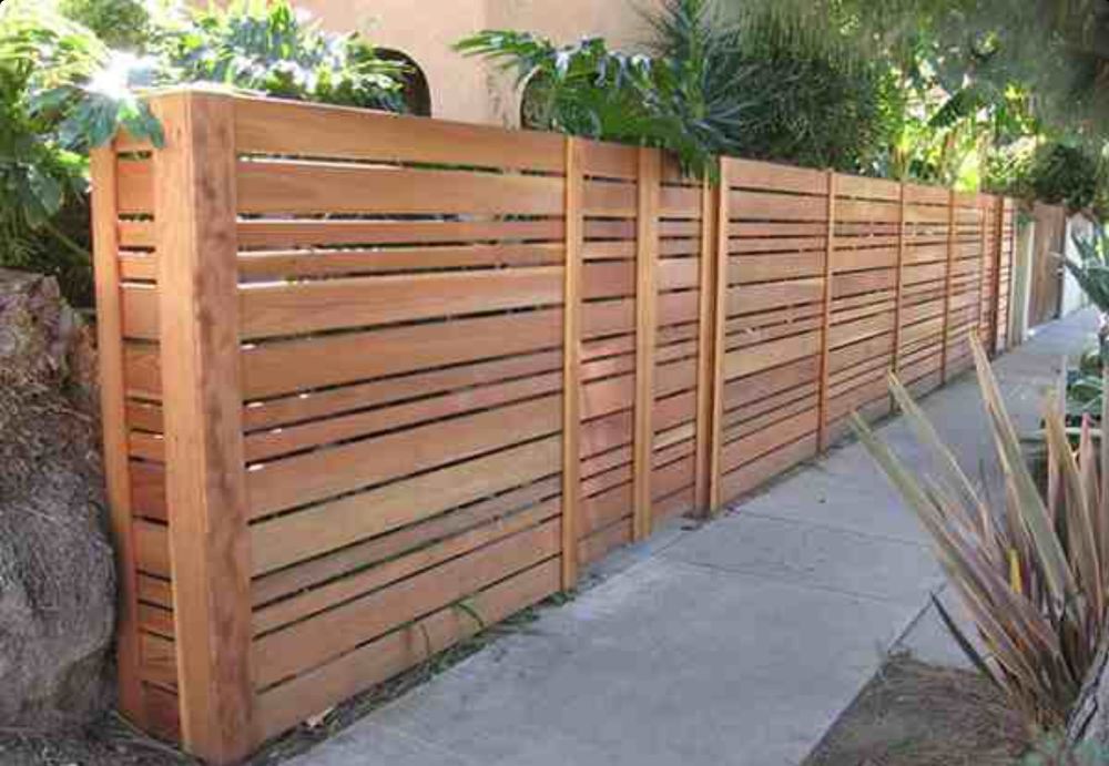 Wooden slat privacy fence along a sidewalk, bordered by greenery and rocks