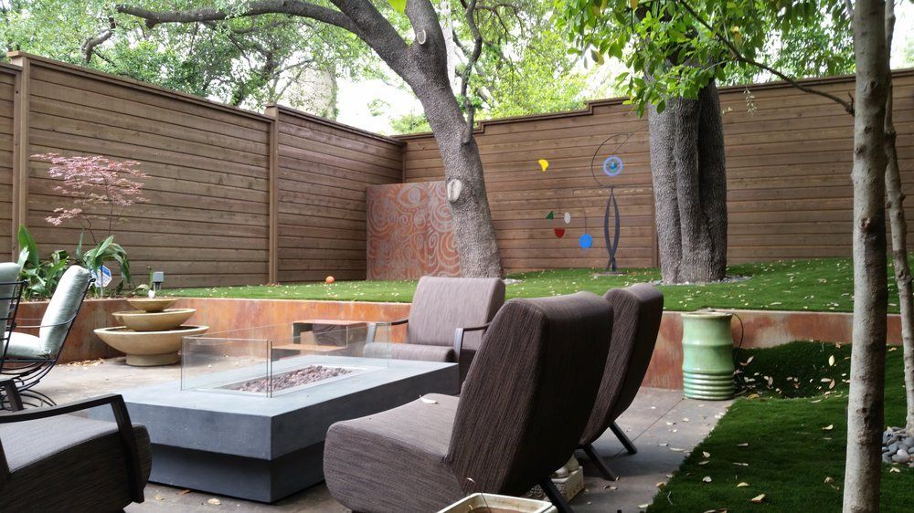 Modern backyard patio with gray seating, fire pit, wooden fence, and large tree over gravel and grass.