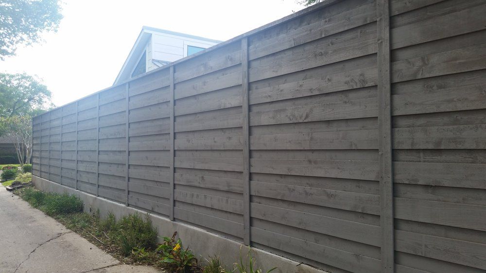 Tall gray wooden privacy fence along a sidewalk beside a residential driveway.