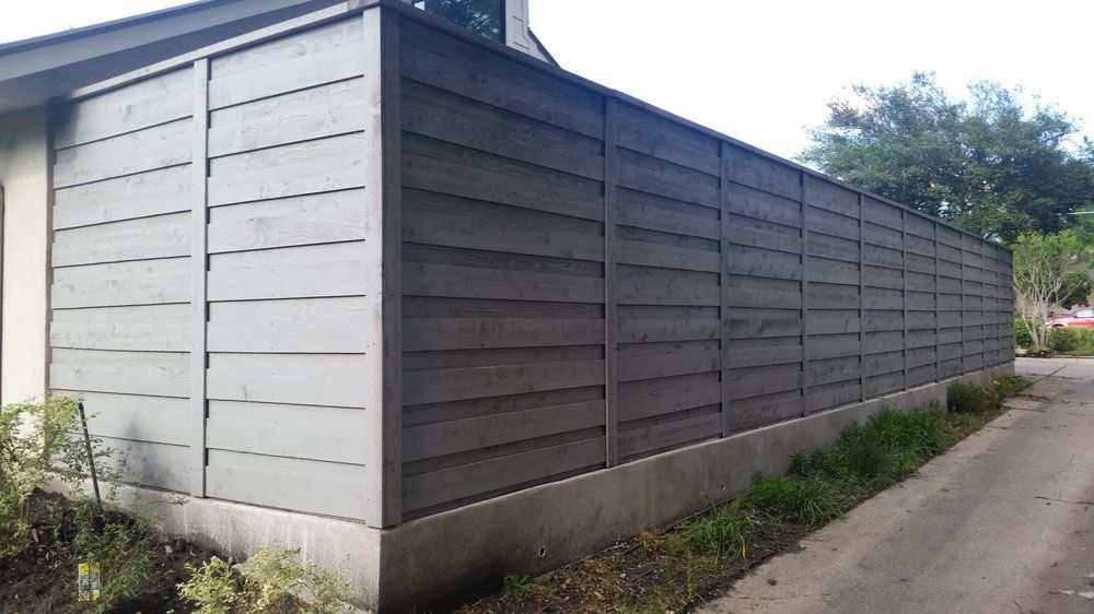 Long gray slatted fence beside a narrow dirt road, with greenery and trees in the background.