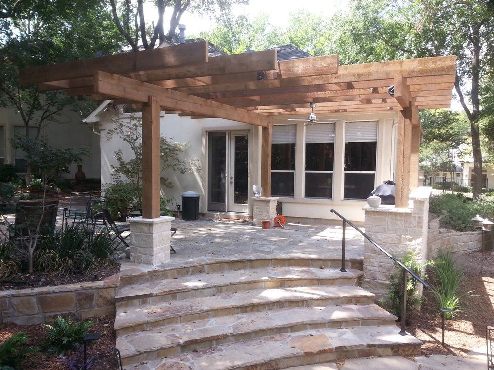 Stone steps lead to a patio with a wooden pergola over a house entrance and windows
