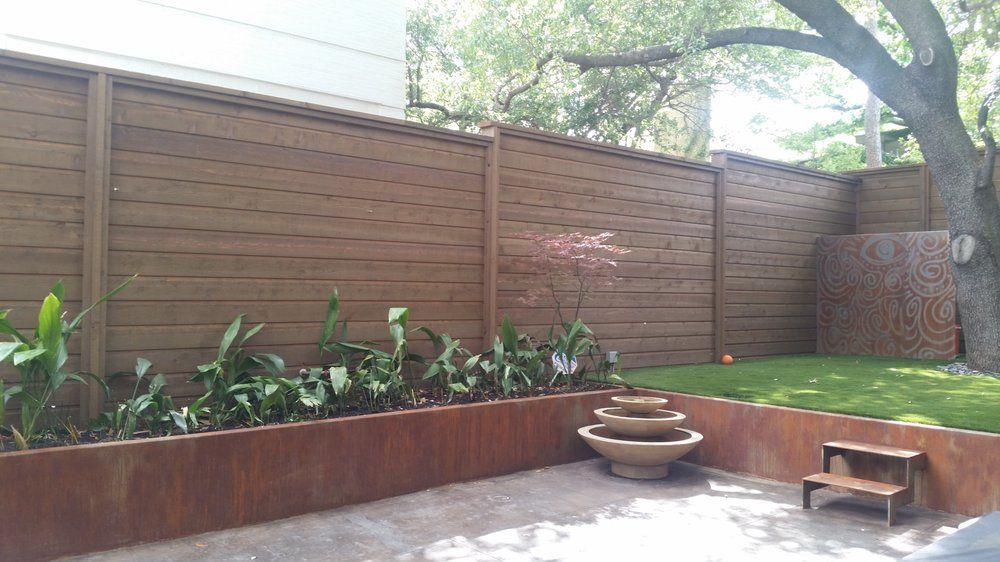 Backyard patio with wooden fence, raised planters, fountain, and grassy corner under trees