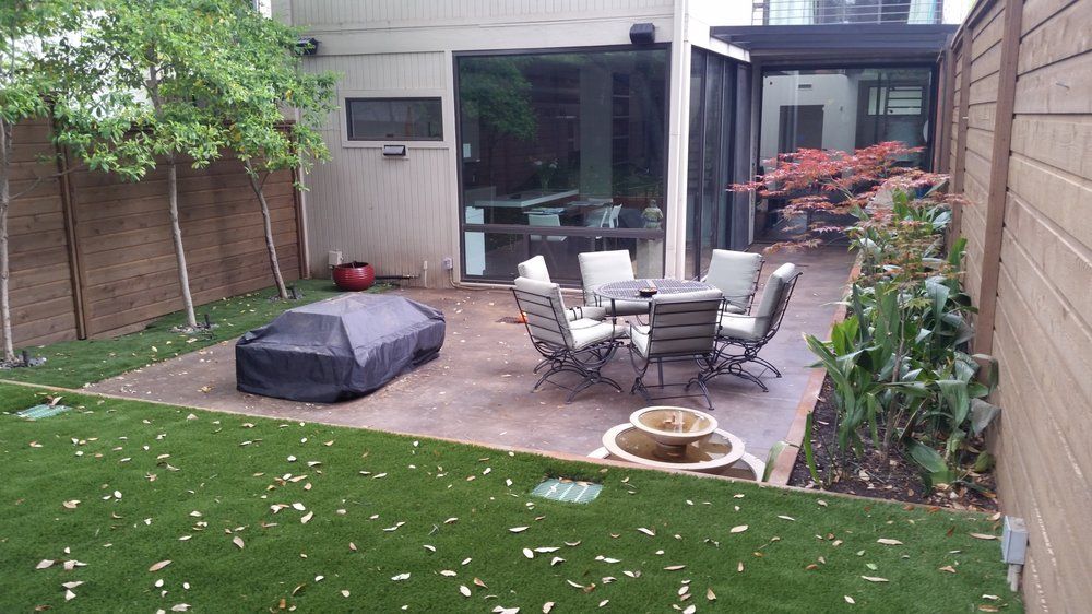 Small backyard patio with table, chairs, grill cover, potted fountain, and surrounding grass and plants.