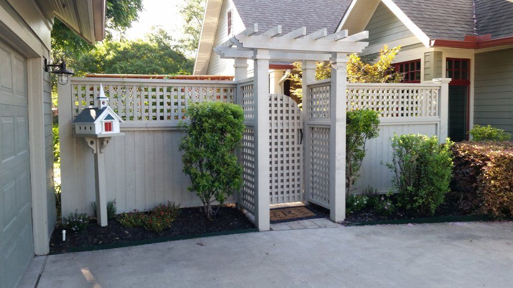 Patio gate with white lattice fencing, small bushes, and a birdhouse beside a house driveway