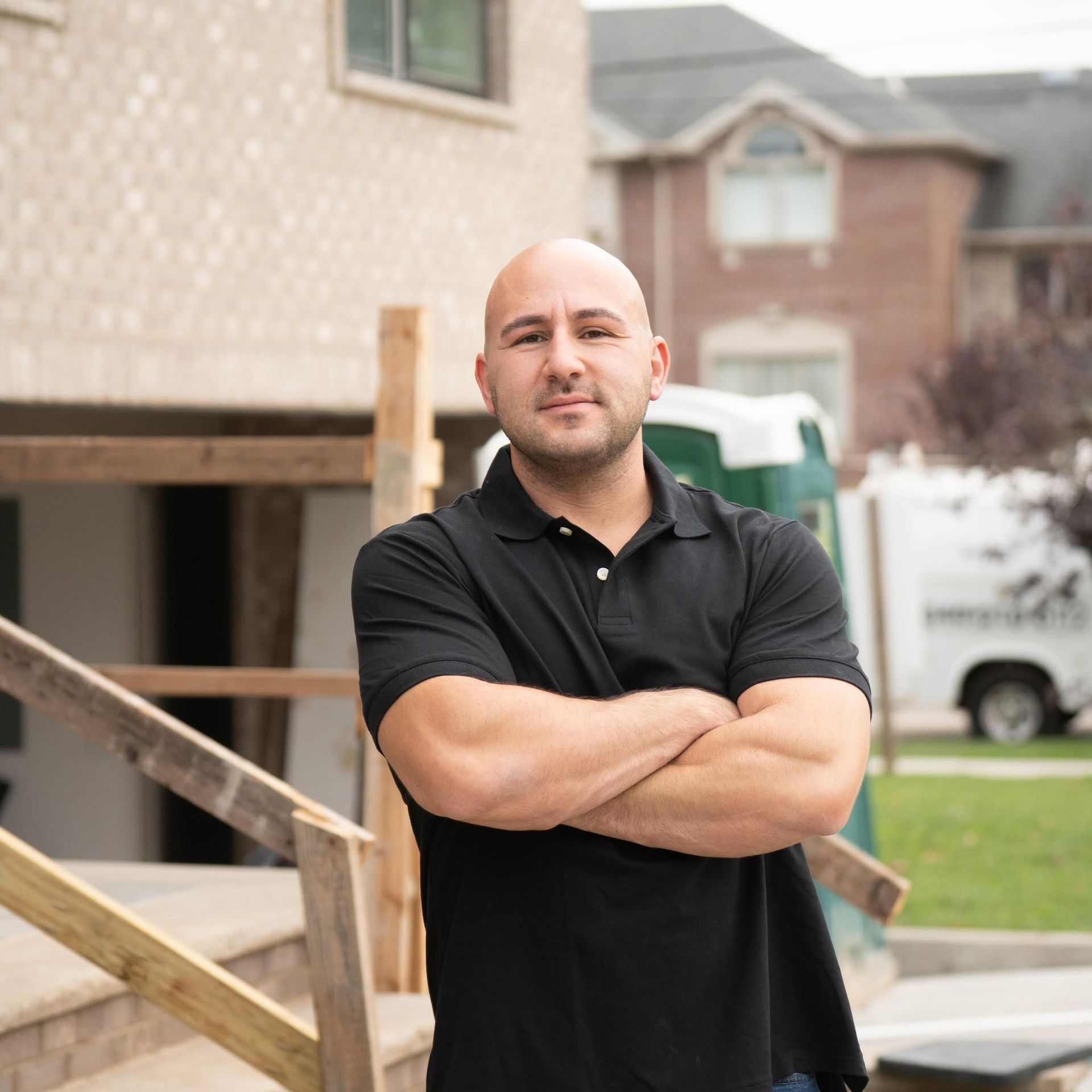 Man standing with arms crossed in front of a house under construction, wearing a black polo shirt.