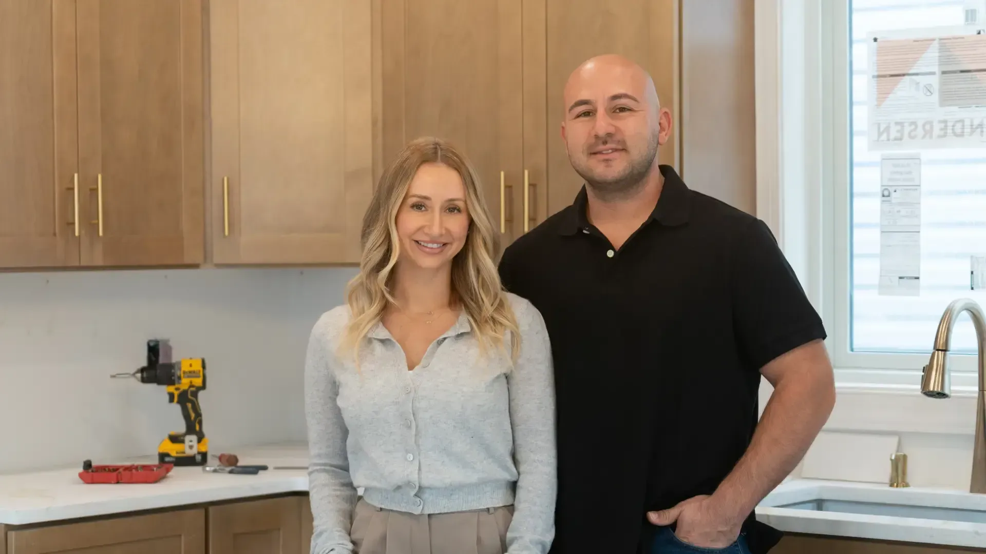 Two people standing in a bright kitchen beside wood cabinets and a white countertop, smiling at the camera