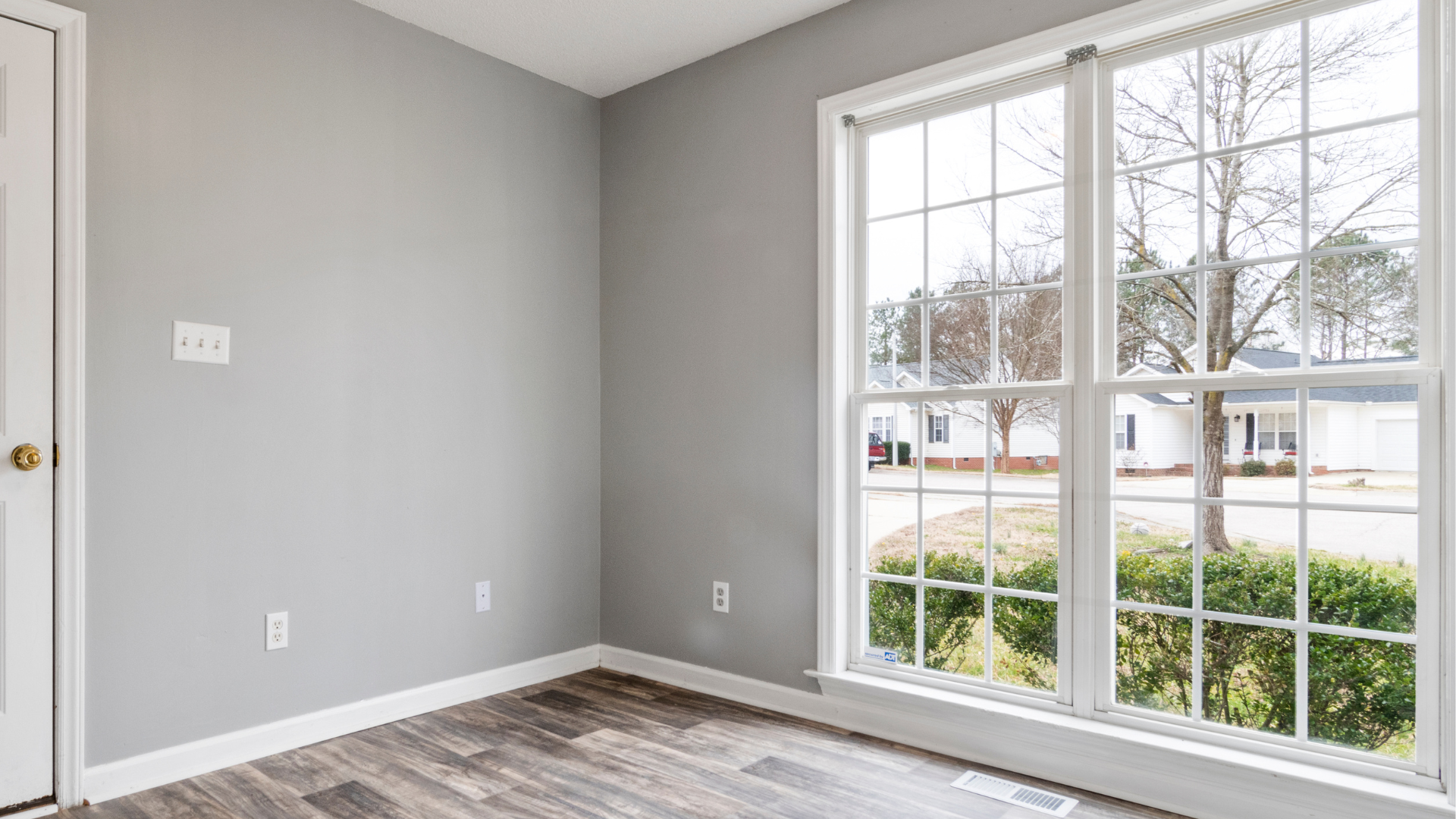 Room with gray walls, wooden floor, and a large window looking onto a suburban street.