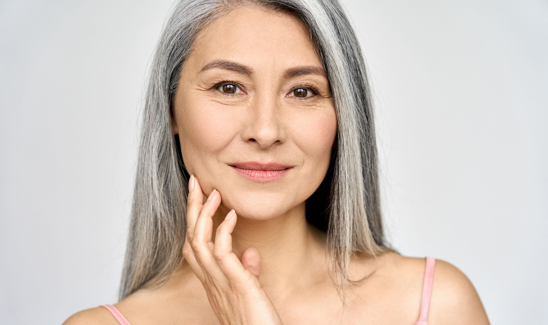 Femme aux longs cheveux gris, souriant doucement en touchant sa joue du bout des doigts, sur un fond gris clair.
