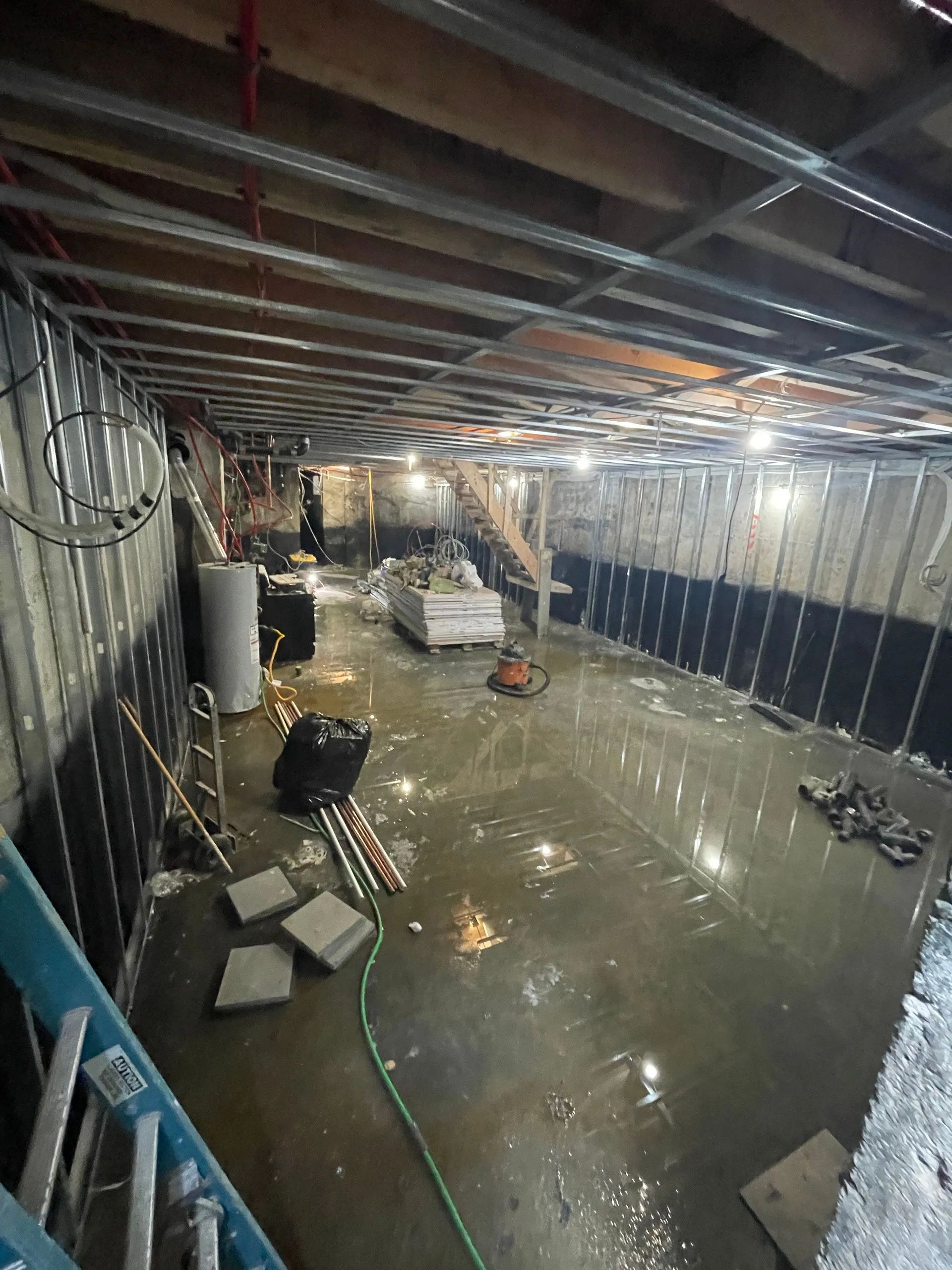 Flooded basement with standing water, exposed beams, hanging lights, and scattered debris.