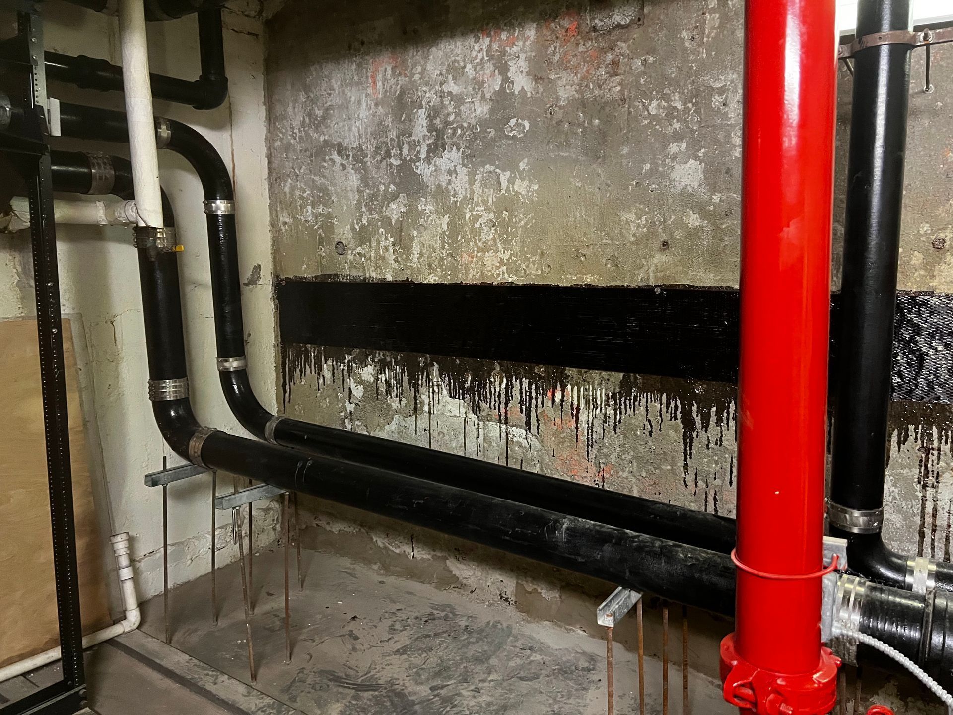 Black pipes and a bright red fire sprinkler pipe run along a textured wall marked with black, dripping waterproof sealant.