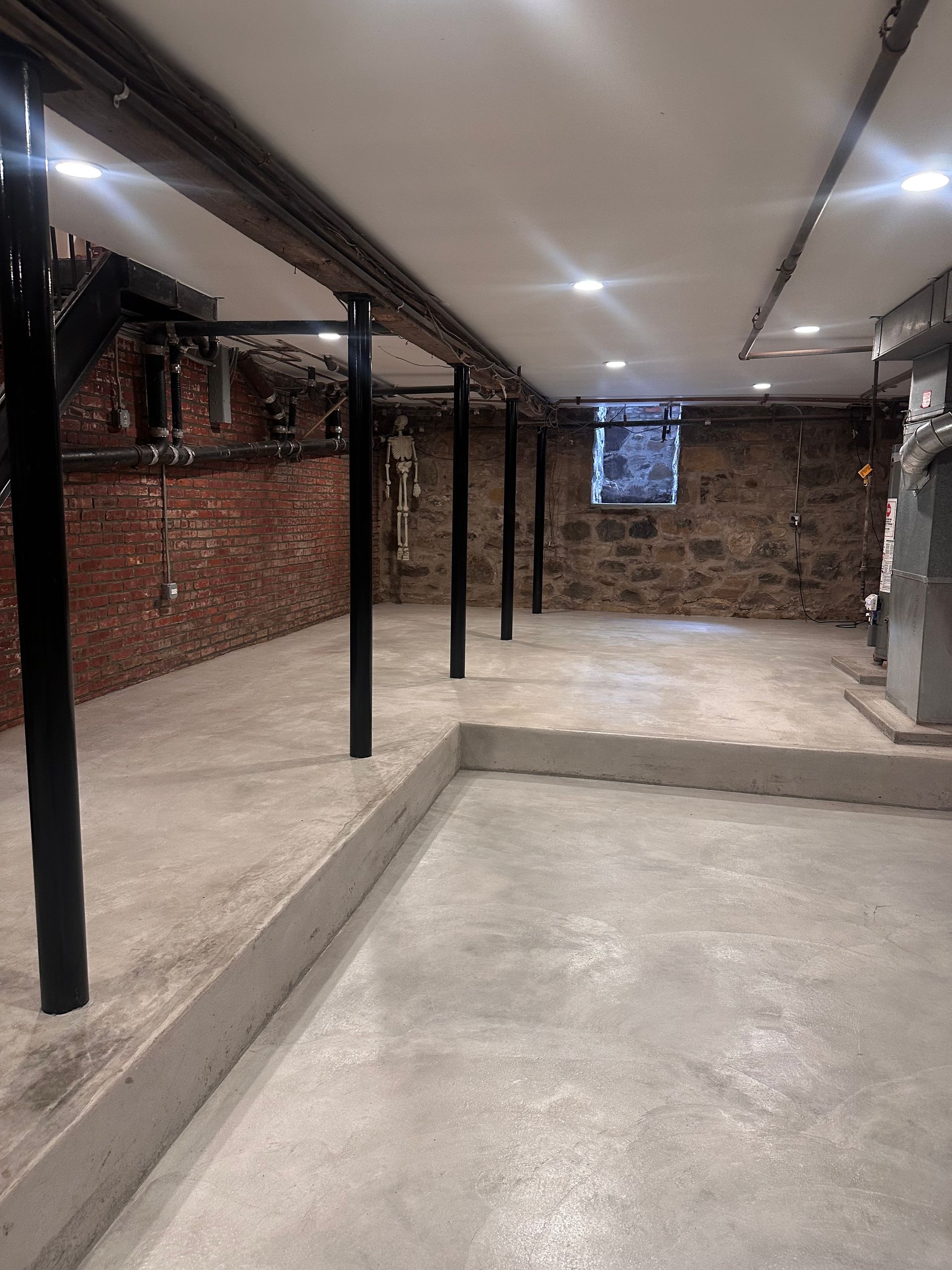 Concrete-floored basement with support beams, exposed brick walls, and stone wall with a small window.