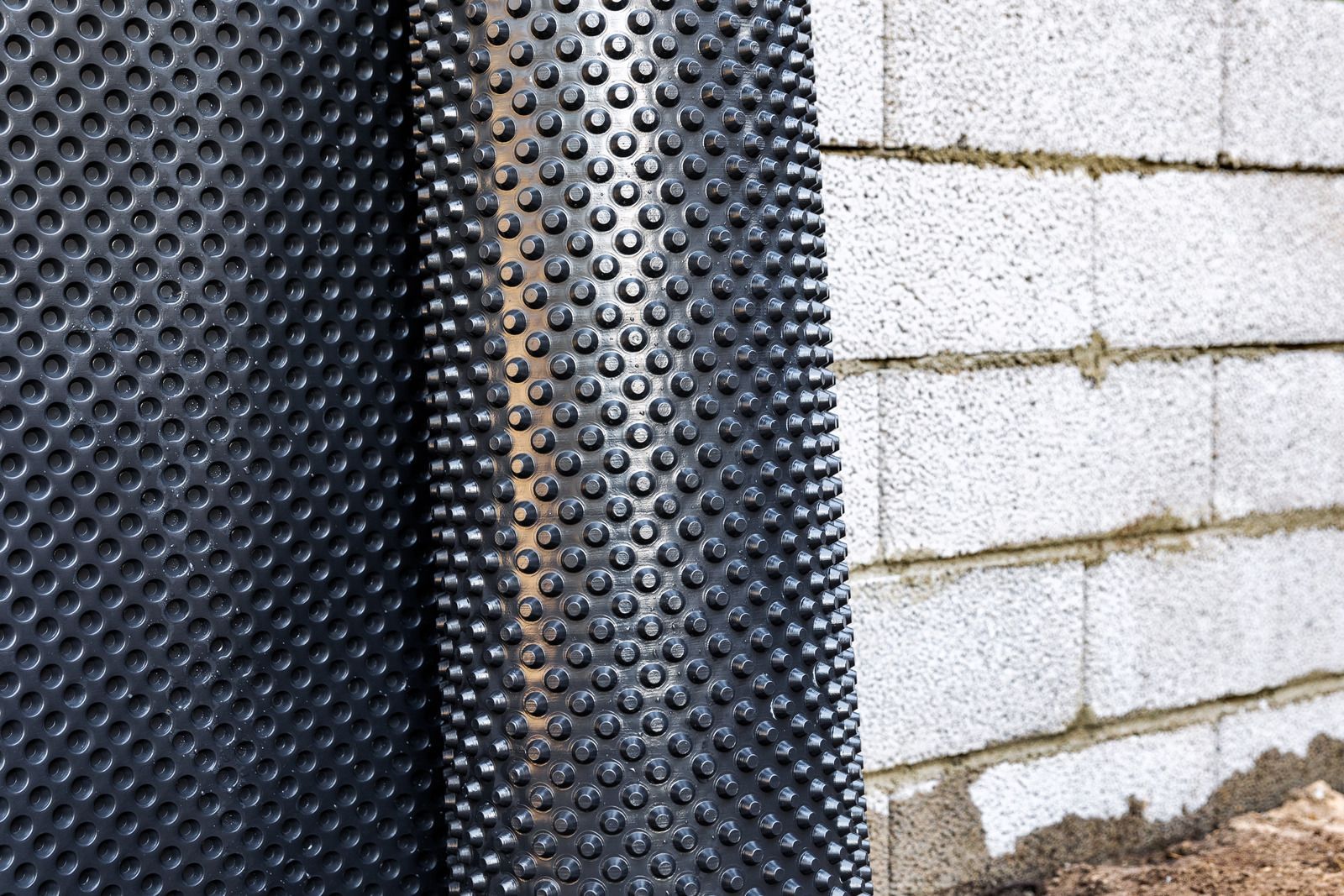 Black dimpled drainage membrane beside a white cinderblock wall outdoors