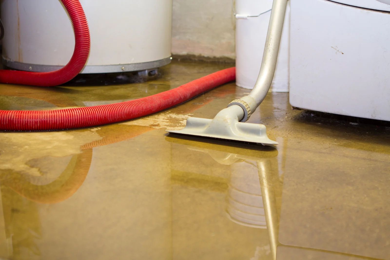Wet floor being cleaned with a vacuum hose and squeegee; red hose beside white machines.