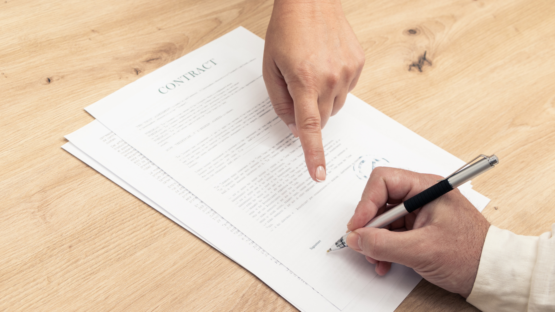 A person is signing a contract with a pen on a wooden table.