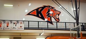 Indoor gym wall with a large orange-and-black wildcat logo and banners above the bleachers