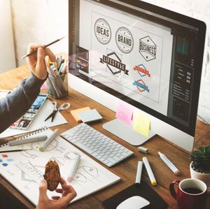 Designer sketching branding ideas at a desk with computer mockups, pens, notes, and coffee