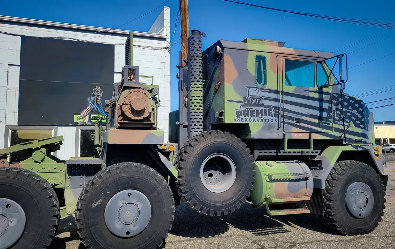 Camouflaged military truck with large off-road tires parked beside a building under a clear blue sky