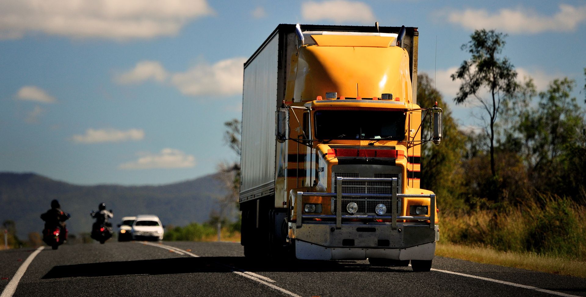 Yellow Semi-truck On A Road With Motorcycles, A Car, And A Mountain In The Background Under A Blue Sky — BA Auto Electrical In West Mackay, QLD