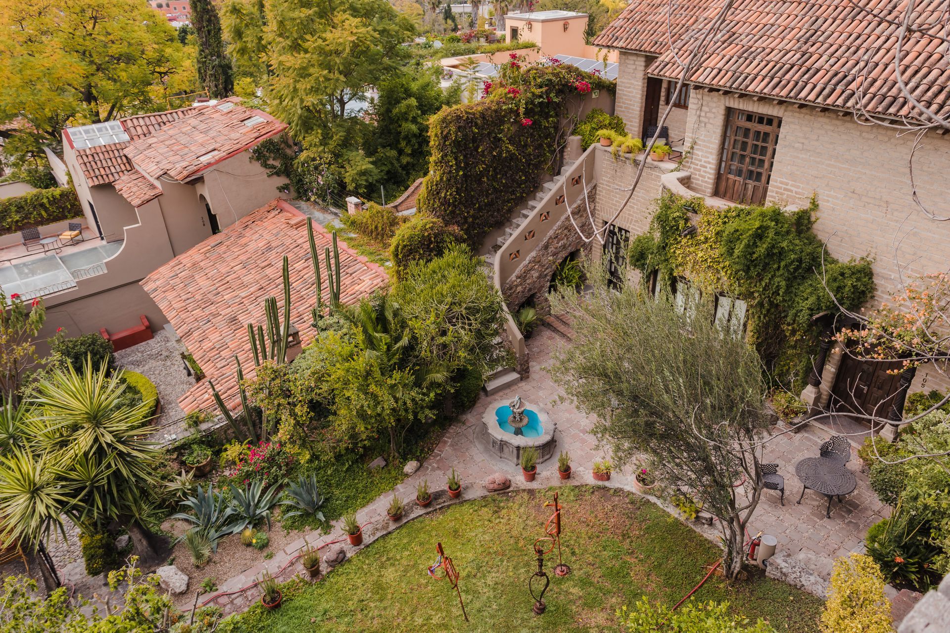 Patio con techos de terracota, exuberante vegetación, fuente, muros de piedra y un camino sinuoso.