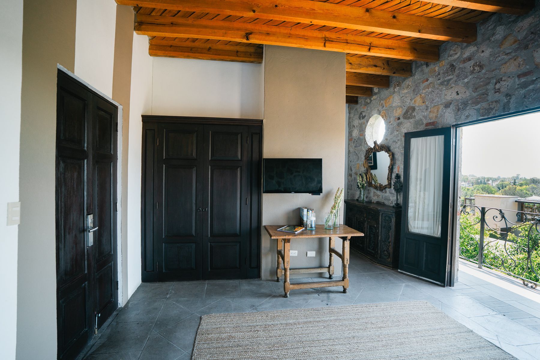 Interior con vigas de madera, pared de piedra, puertas oscuras, mesa pequeña, alfombra y puerta abierta al balcón.