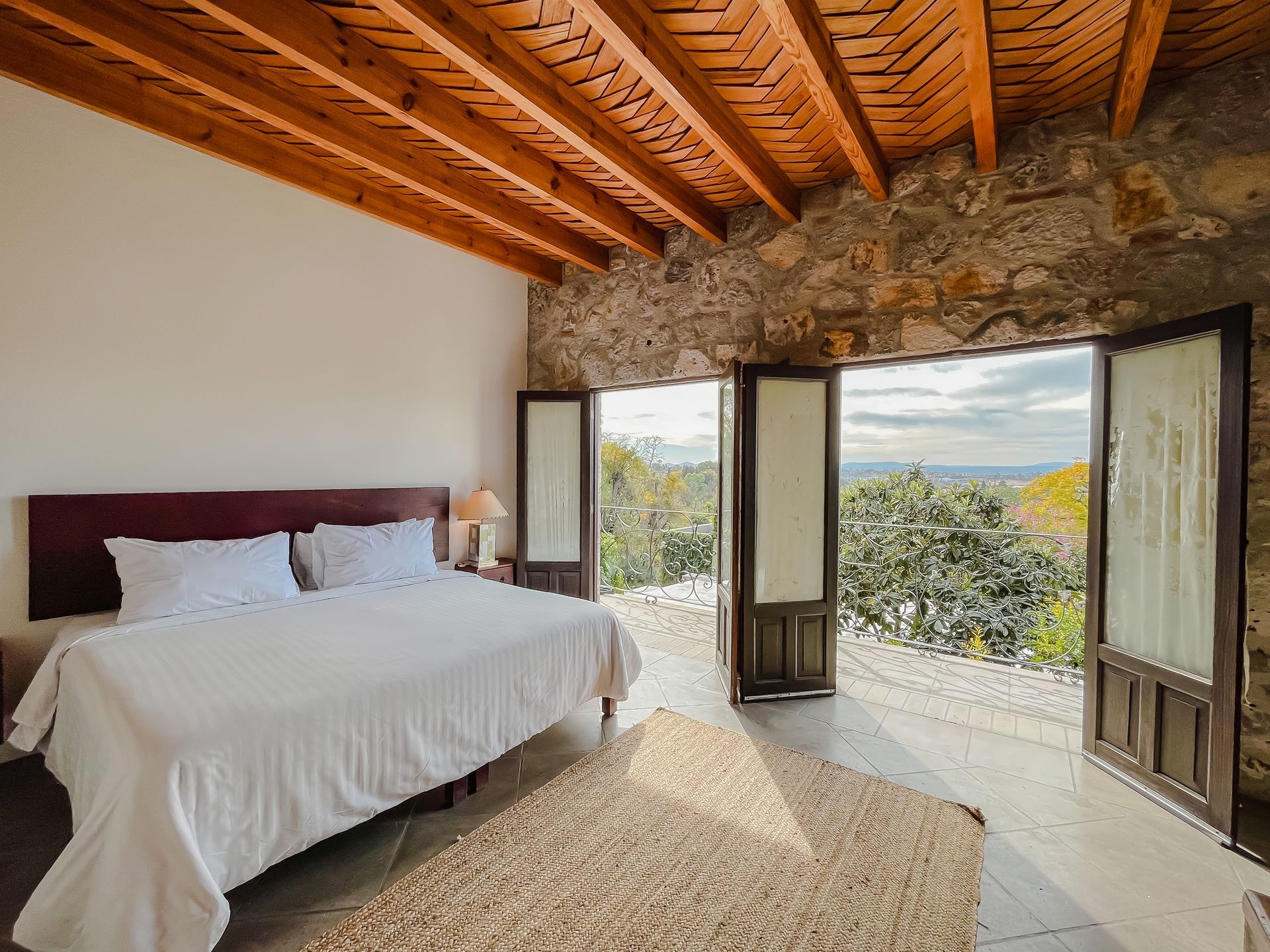 Dormitorio con puertas abiertas y vistas. Pared de piedra y techo de madera. Ropa de cama blanca y alfombra tejida.