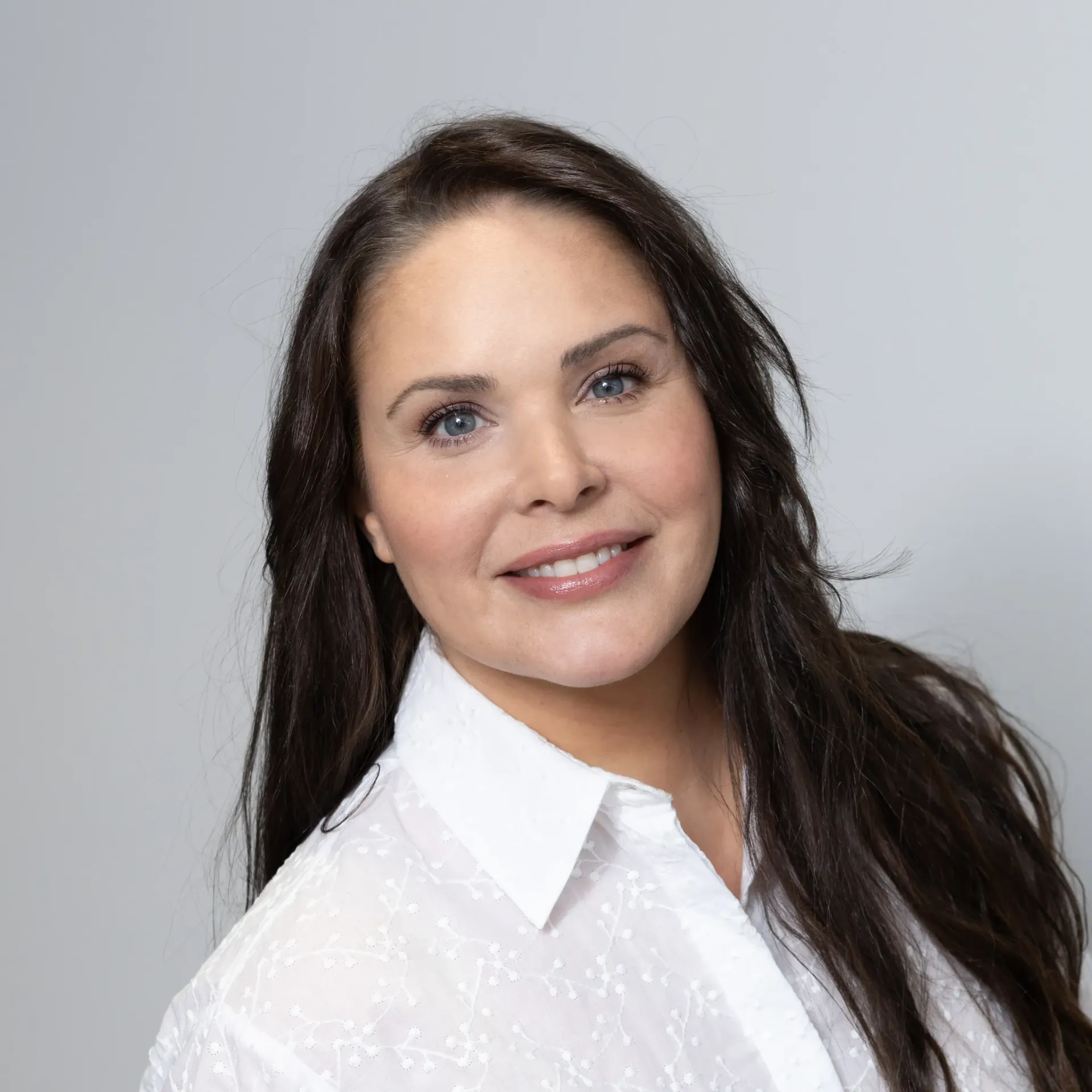 A woman with long dark hair is wearing a white shirt and smiling.