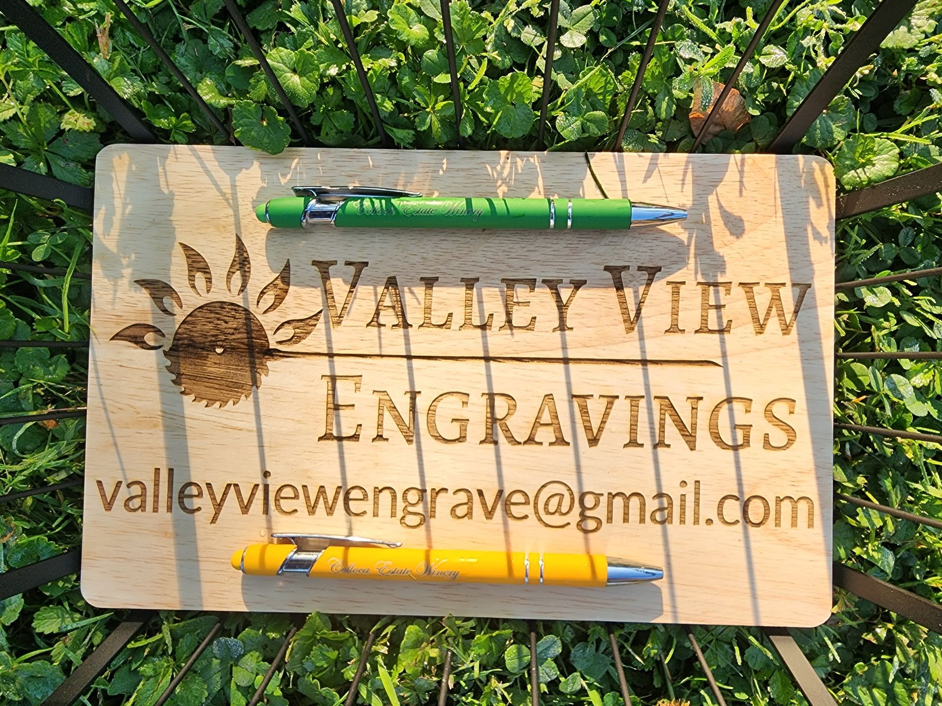 Wooden sign for Valley View Engravings with pens on top, set against green foliage.