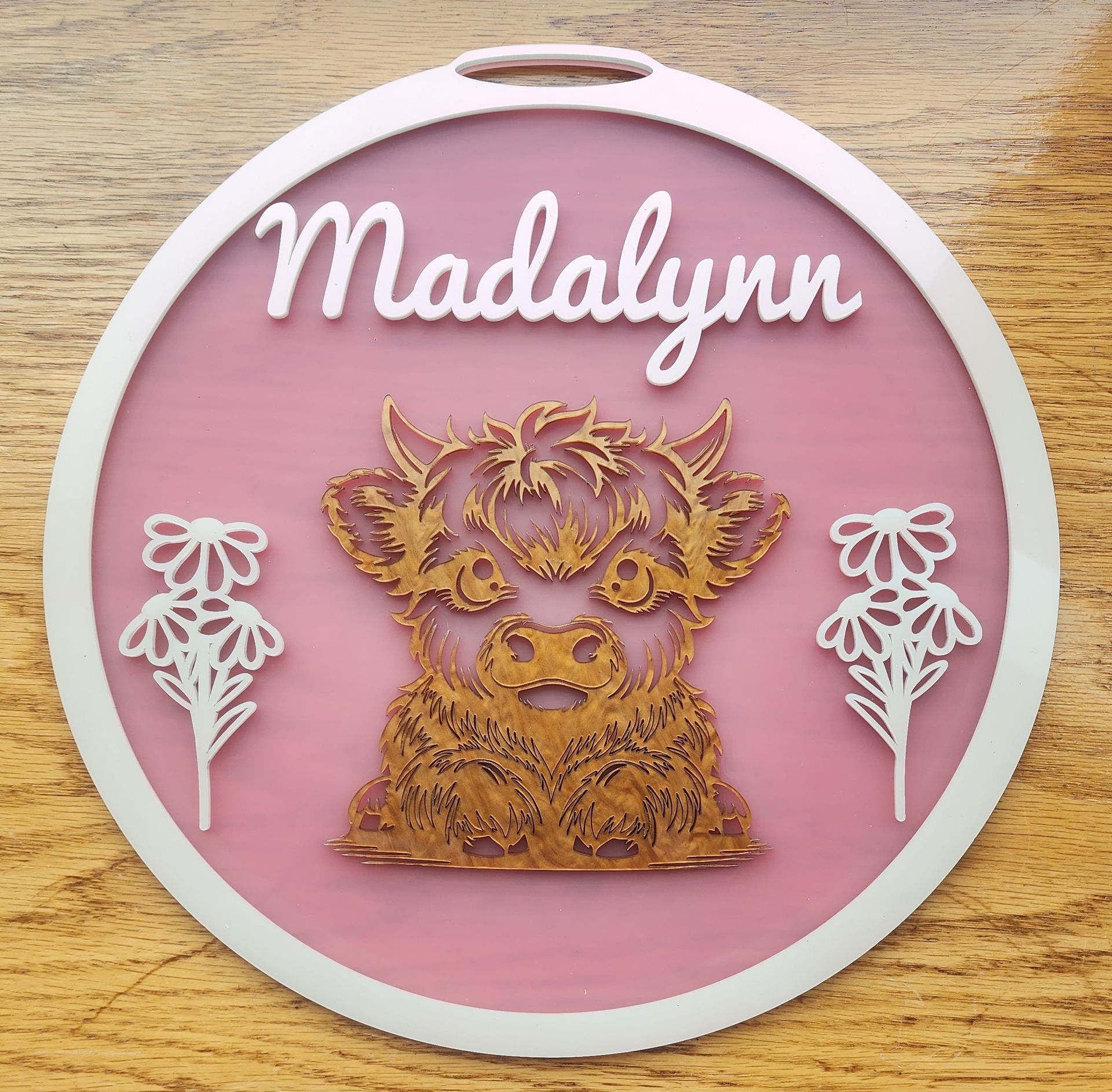 Pink round plaque with “Madalynn” and a golden floral cow illustration on a wood background