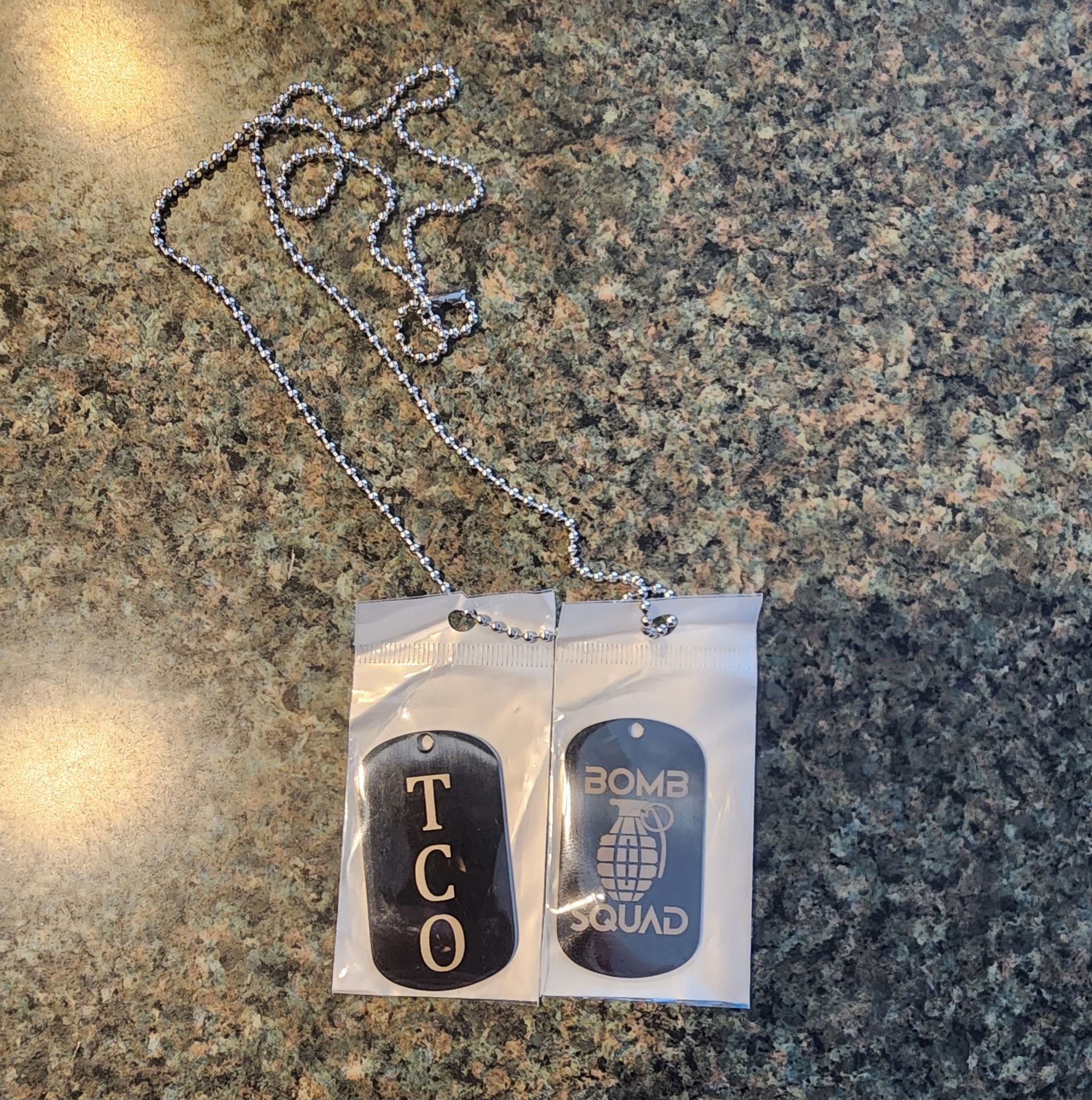 Silver chain necklace with a clear pendant reading “TCO” and “BOMB SQUAD” on a speckled stone surface