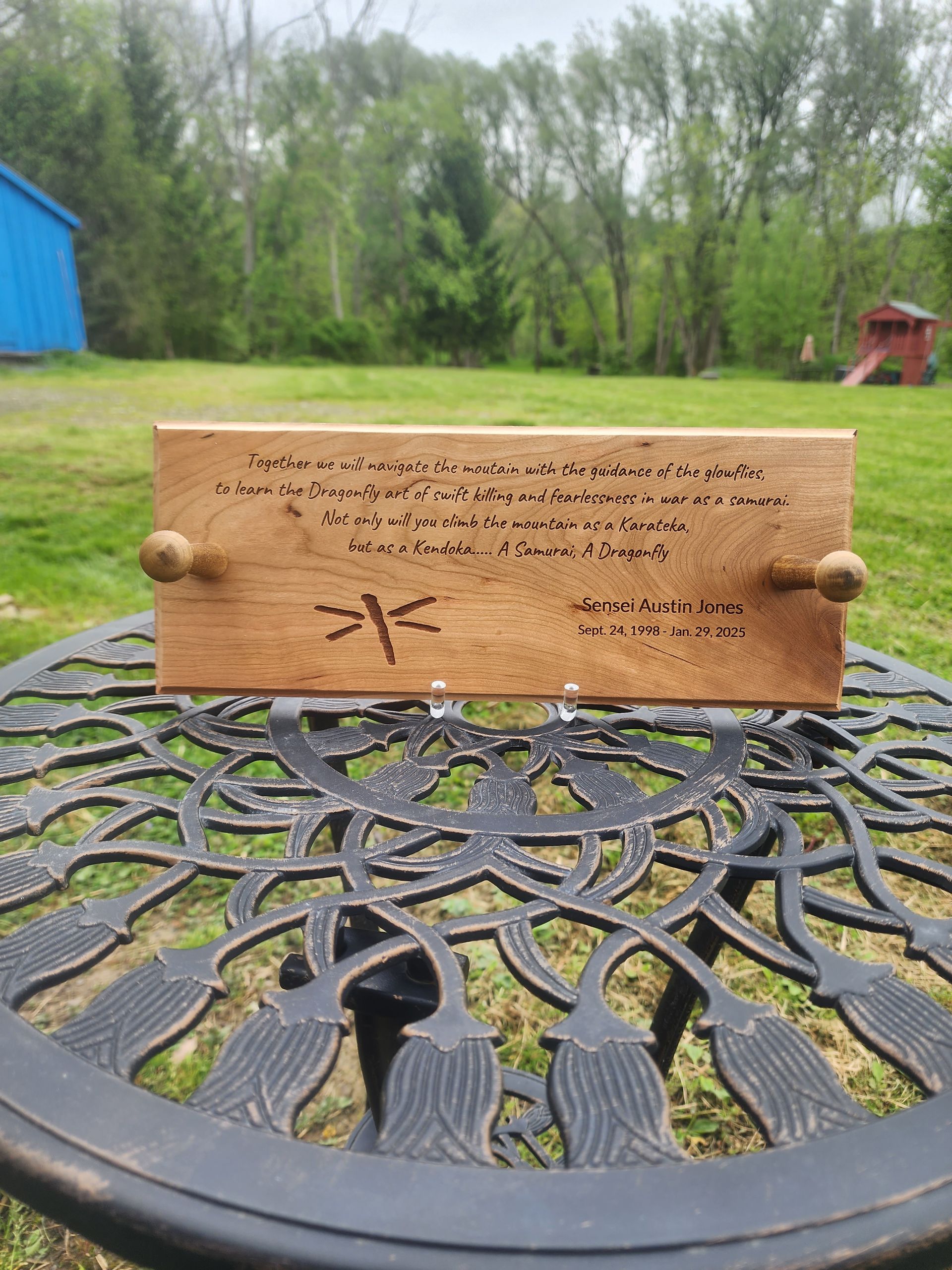 Wooden plaque with engraved text on a black metal table in a grassy yard near trees and a blue shed