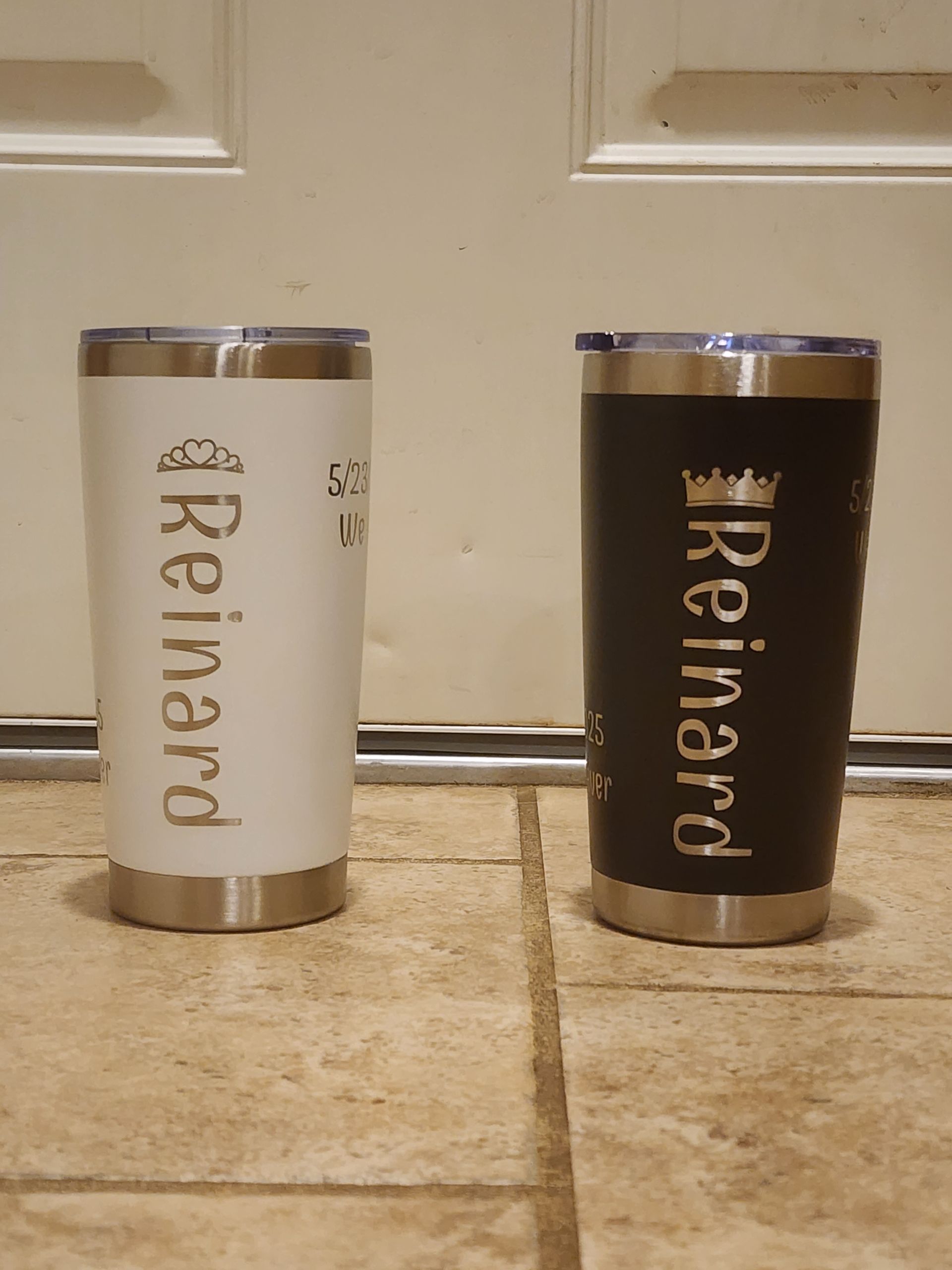 Two custom tumblers, one white and one black, standing on a tiled floor in front of a door.