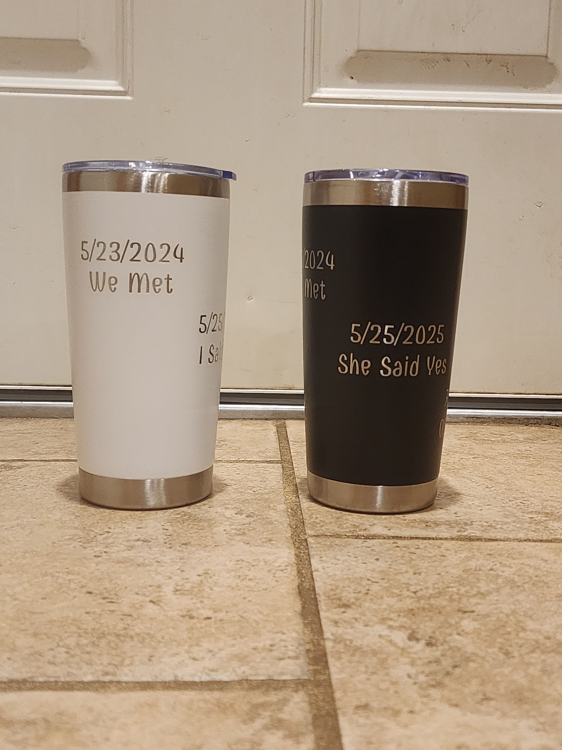 Two stainless steel tumblers on a tiled floor, one white and one black, both with printed dates.