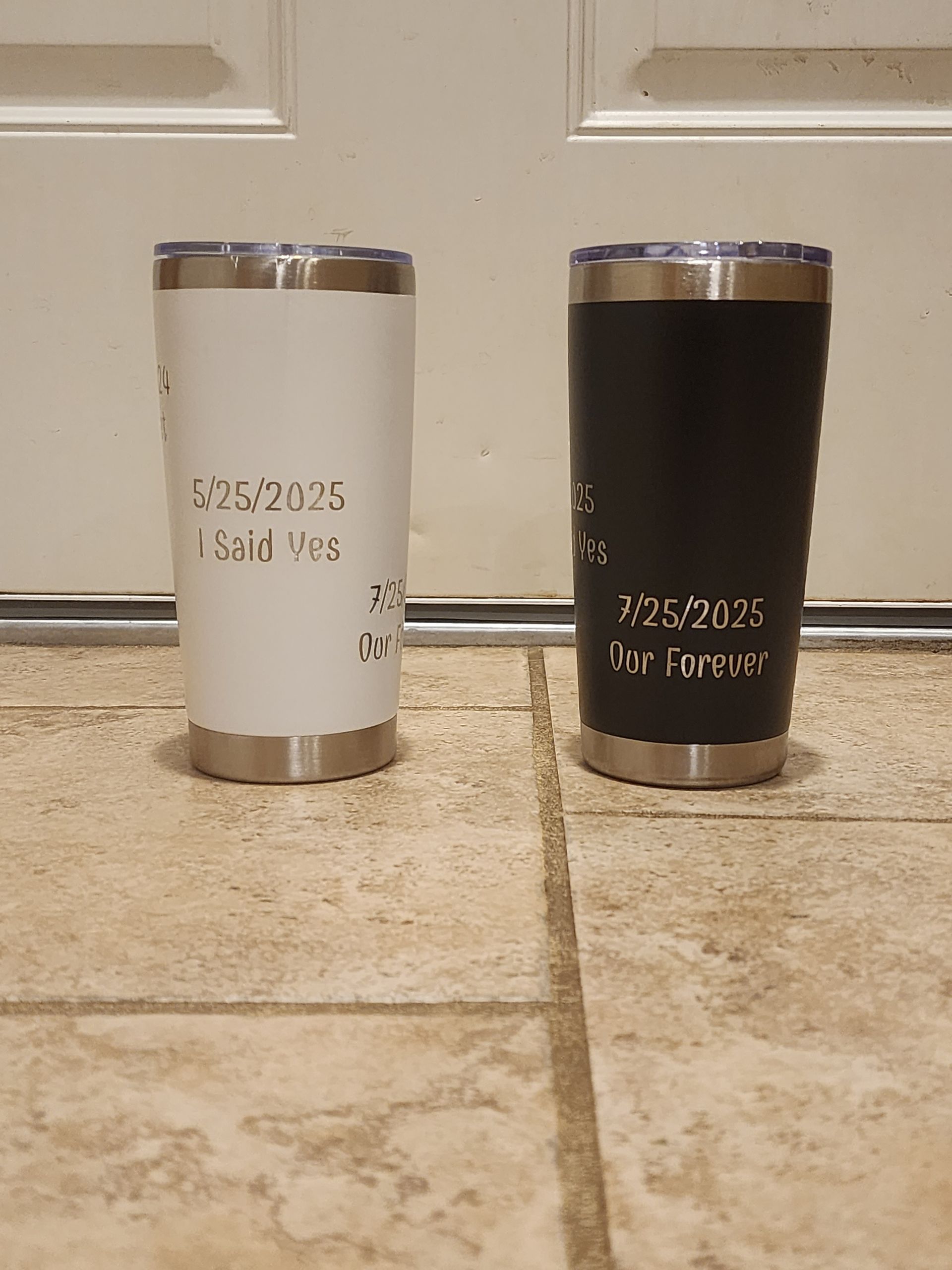 Two stainless steel tumblers, one white and one black, standing on a tiled floor against a wall.