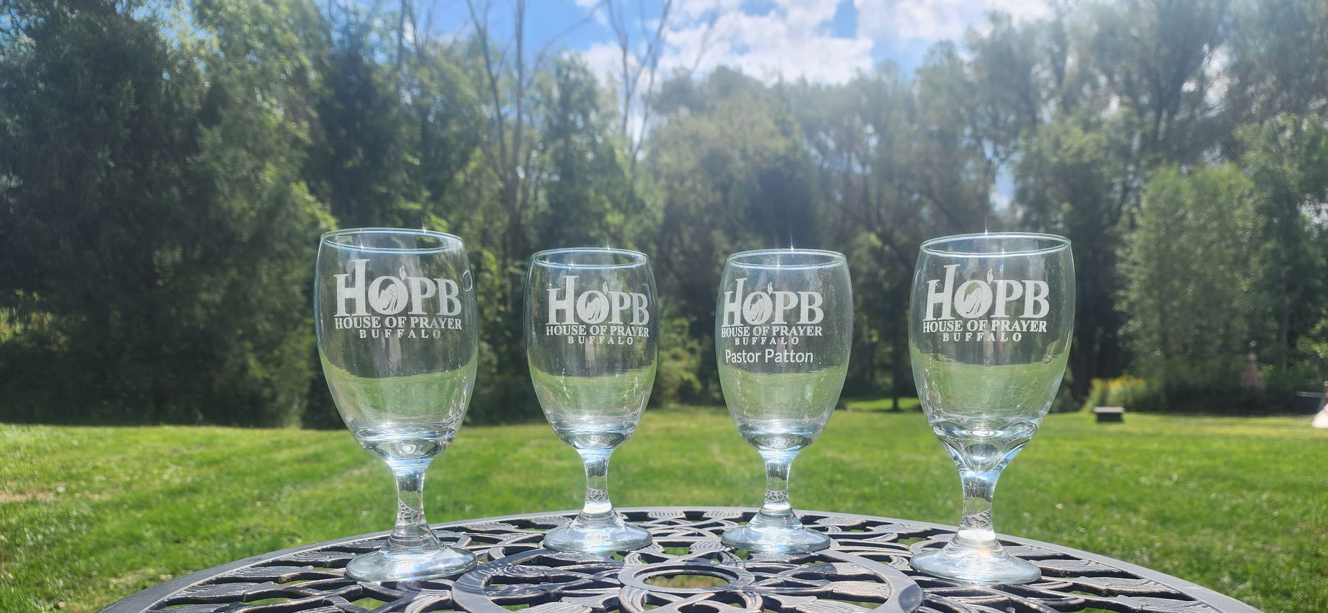 Four etched HOP glasses on a patio table, with a sunny green lawn and trees in the background.