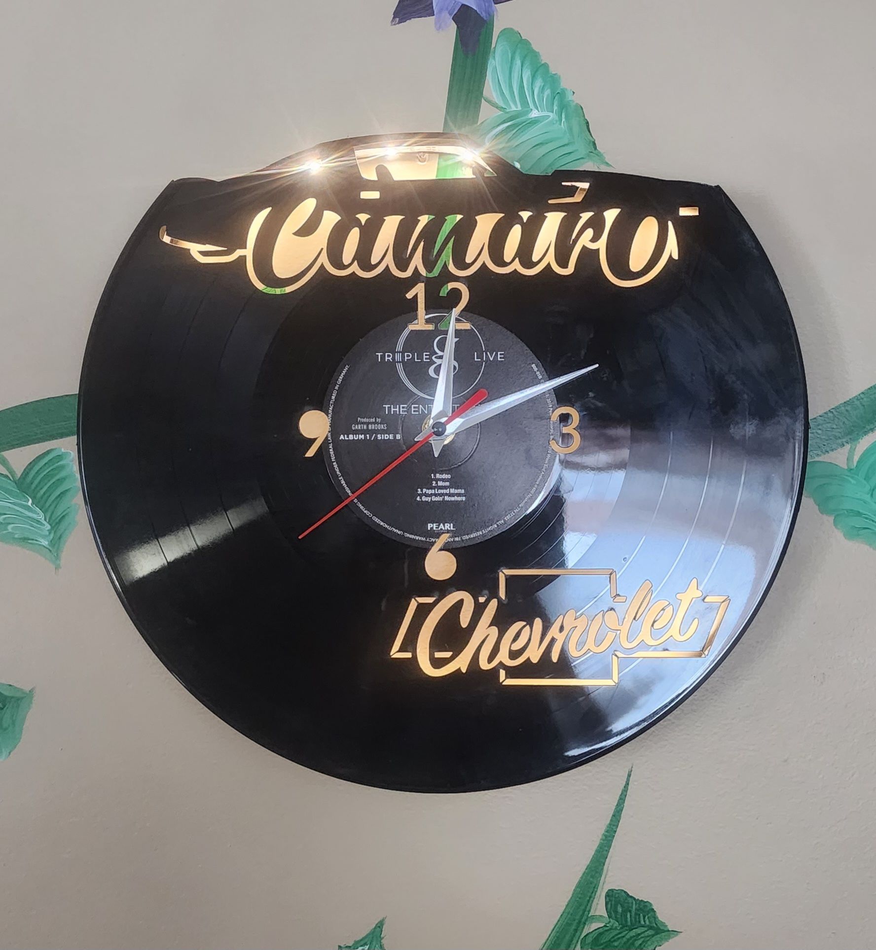 Black wall clock with gold lettering and leaf decorations on a white wall.