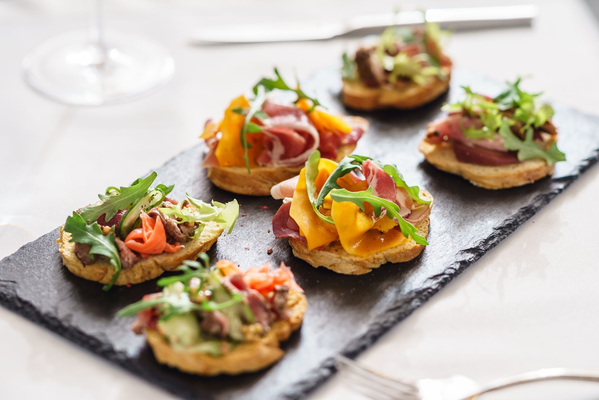 Bruschetta-Häppchen mit verschiedenen Belägen, angerichtet auf einer schwarzen Schieferplatte, weißes Gedeck.