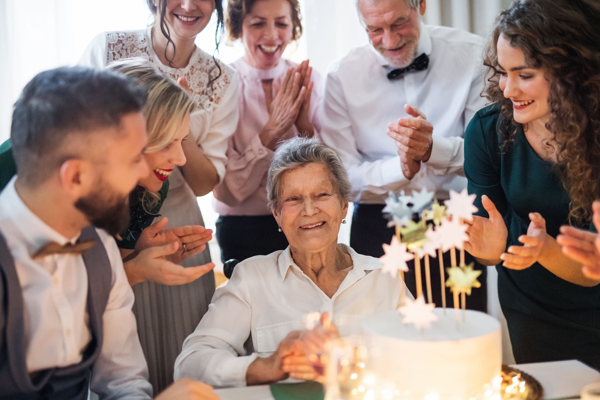 Eine Familie feiert auf einer Geburtstagsfeier; alle sitzen um eine ältere Dame herum, die an einem Tisch mit Kuchen sitzt.