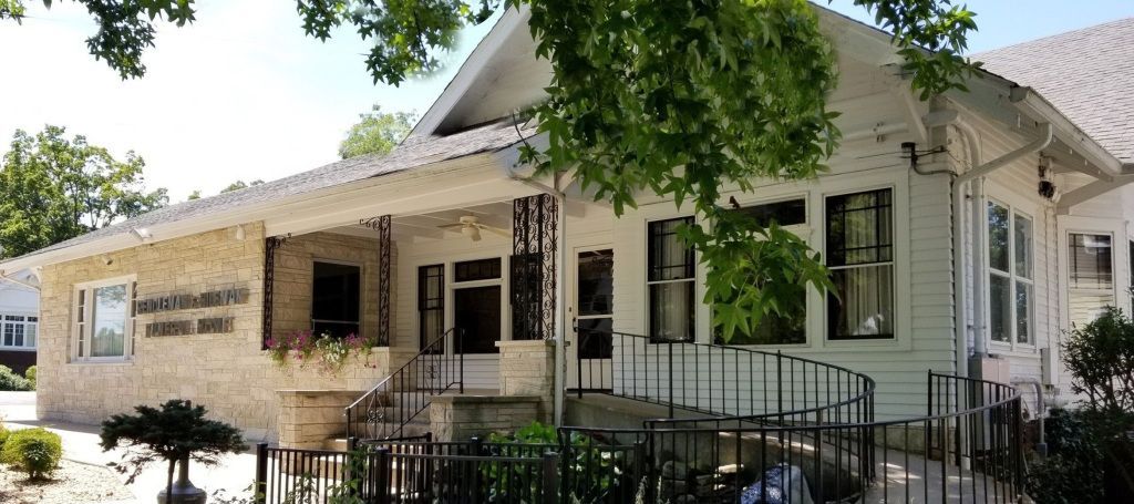 White building with a stone facade, steps, and a ramp. Black railing and trees frame the view.