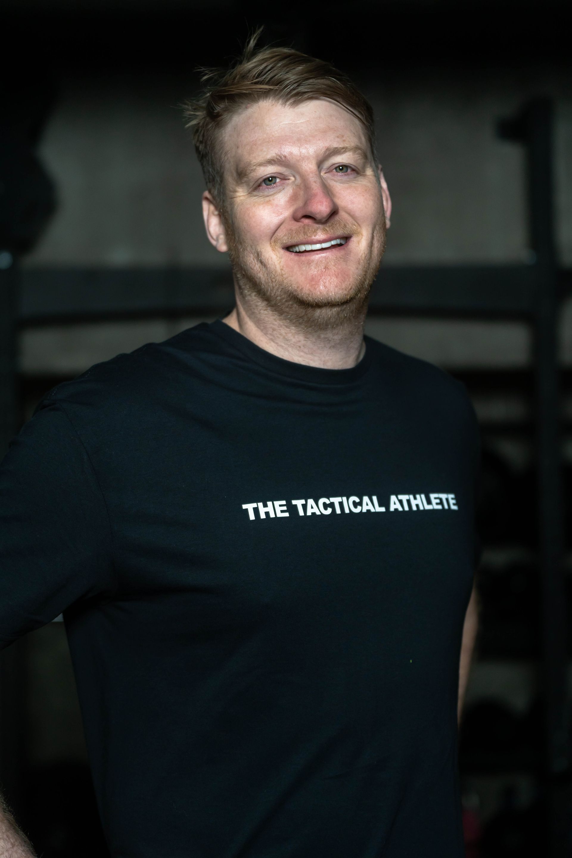 A man wearing a black t-shirt that says `` the tactical athlete '' is smiling.