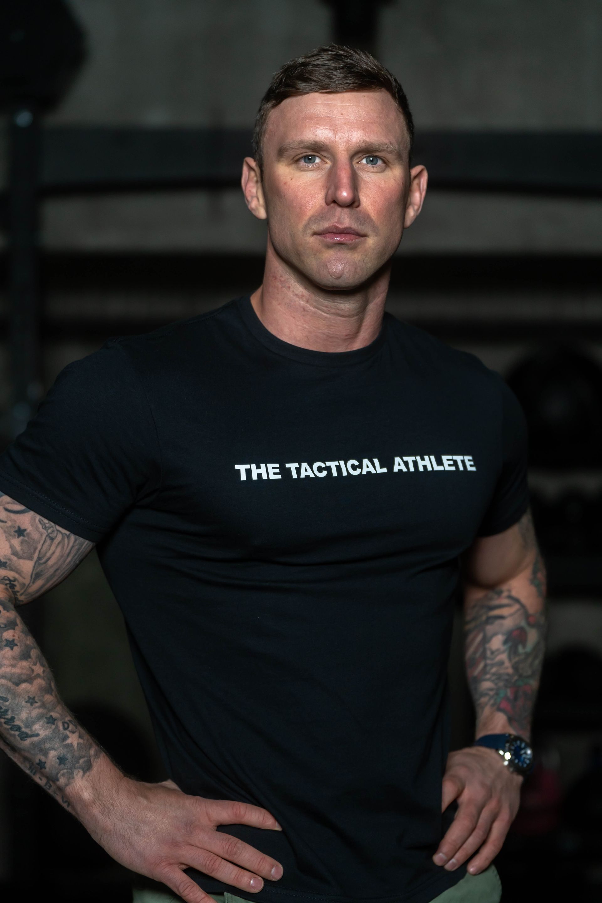 A man wearing a black t-shirt that says the tactical athletes is standing with his hands on his hips.