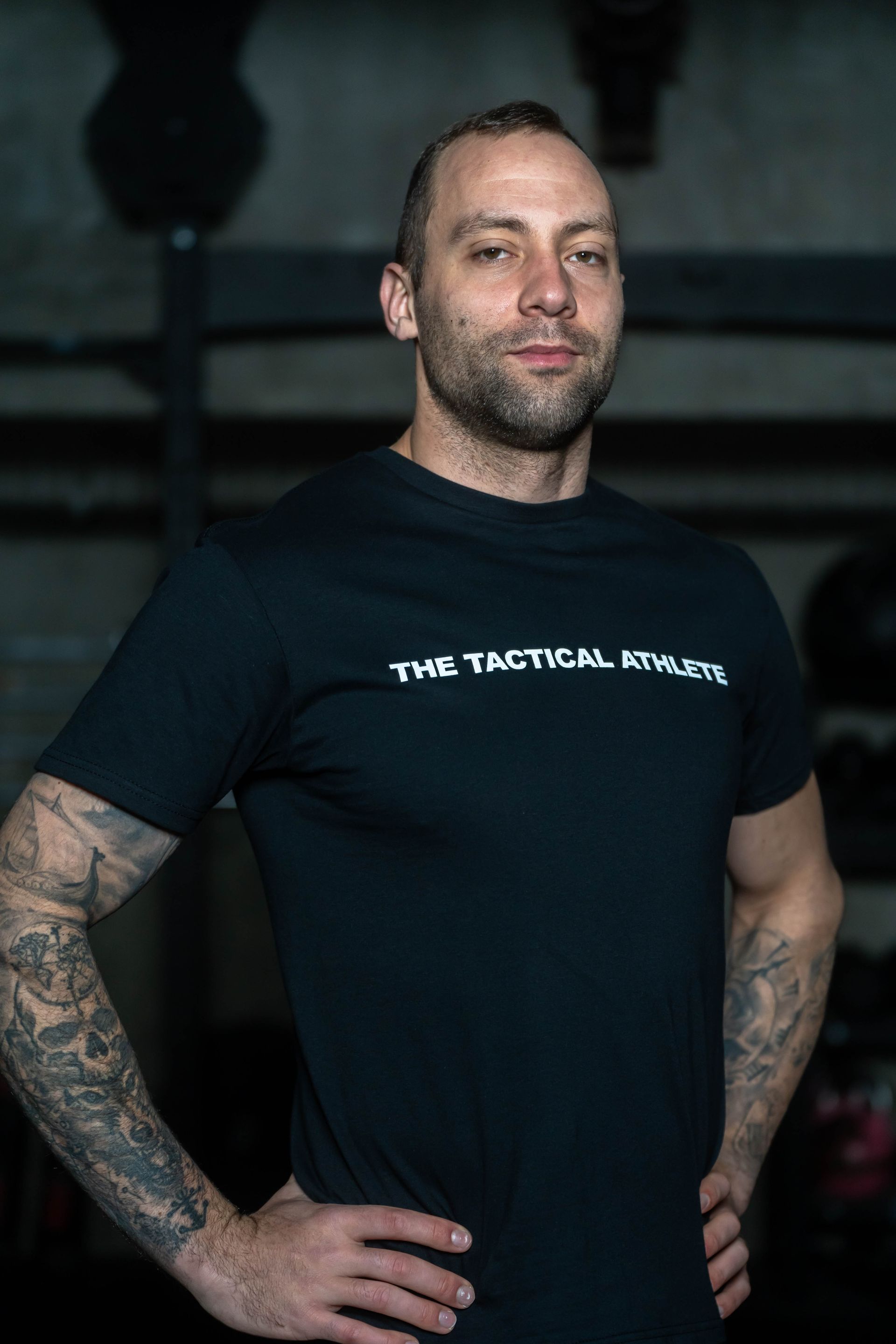 A man with a beard and tattoos is wearing a black t-shirt and standing with his hands on his hips.