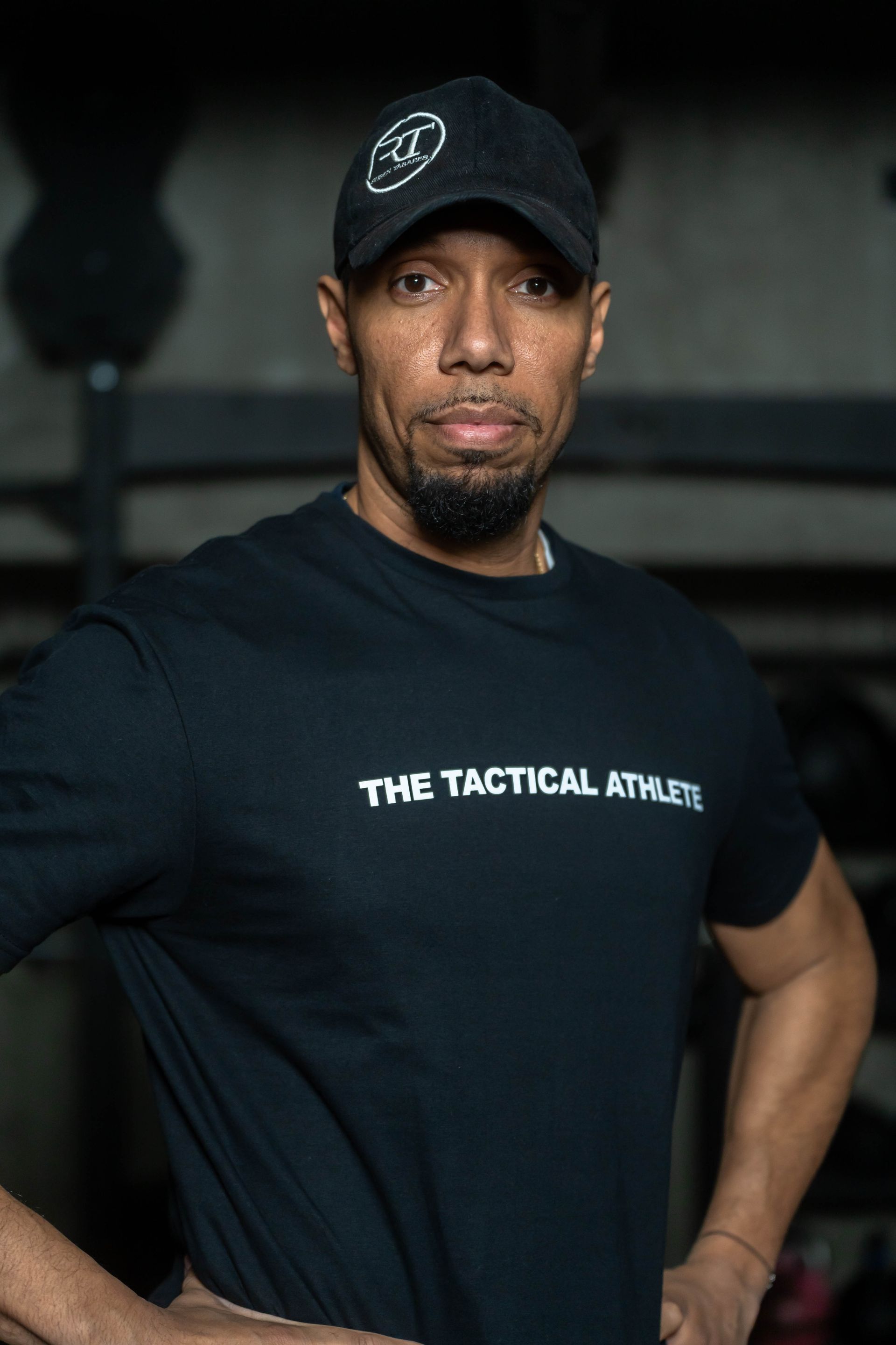 A man wearing a black t-shirt and a hat is standing with his hands on his hips.