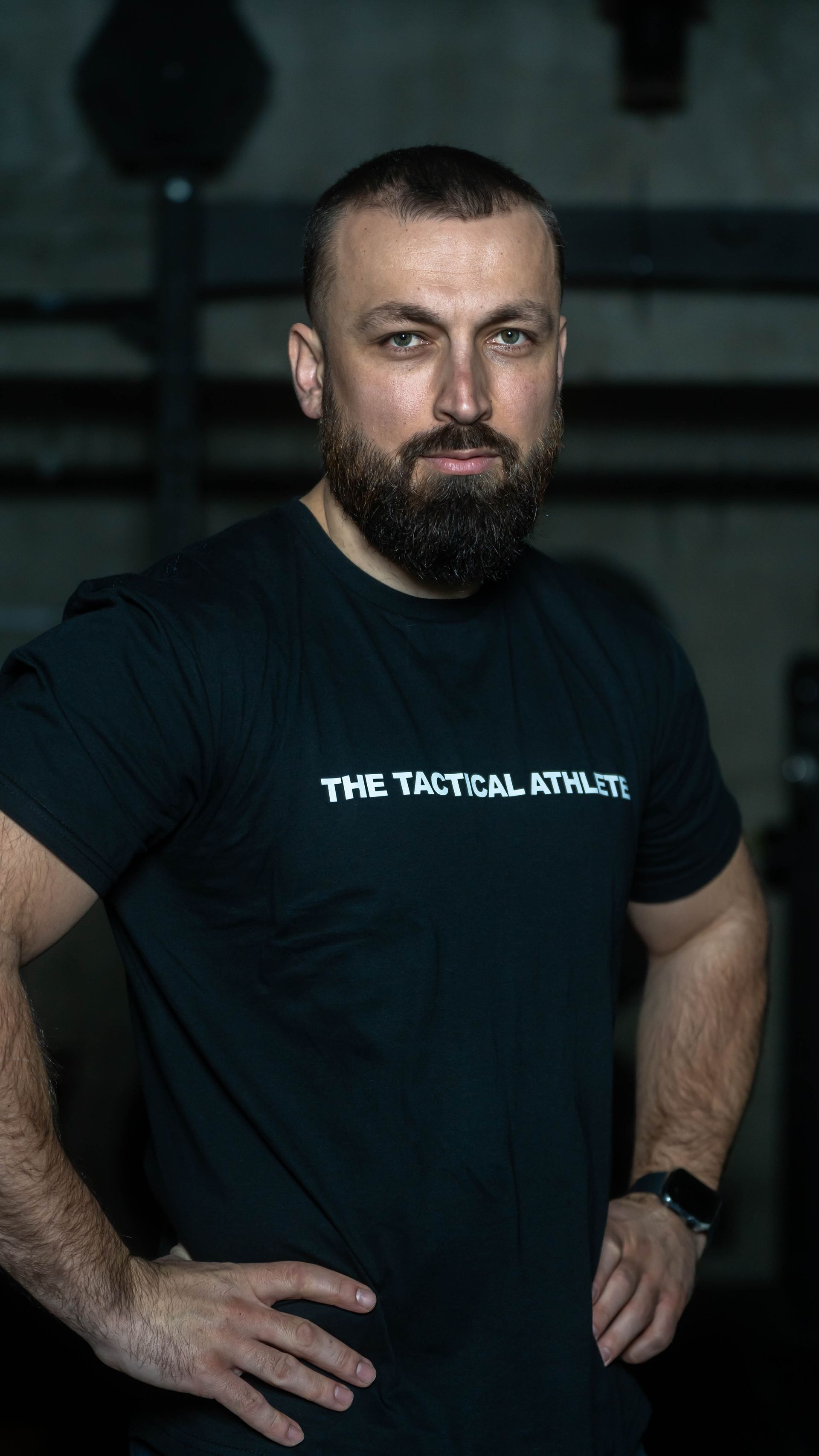 A man with a beard wearing a black t-shirt is standing with his hands on his hips.