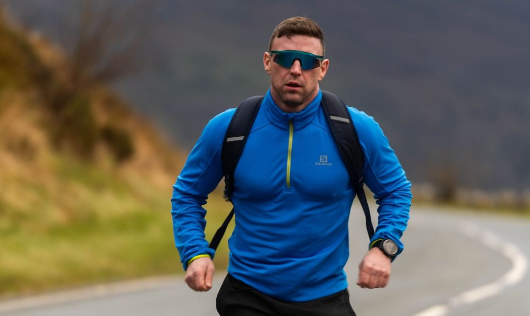 A man wearing sunglasses and a blue shirt is running down a road.