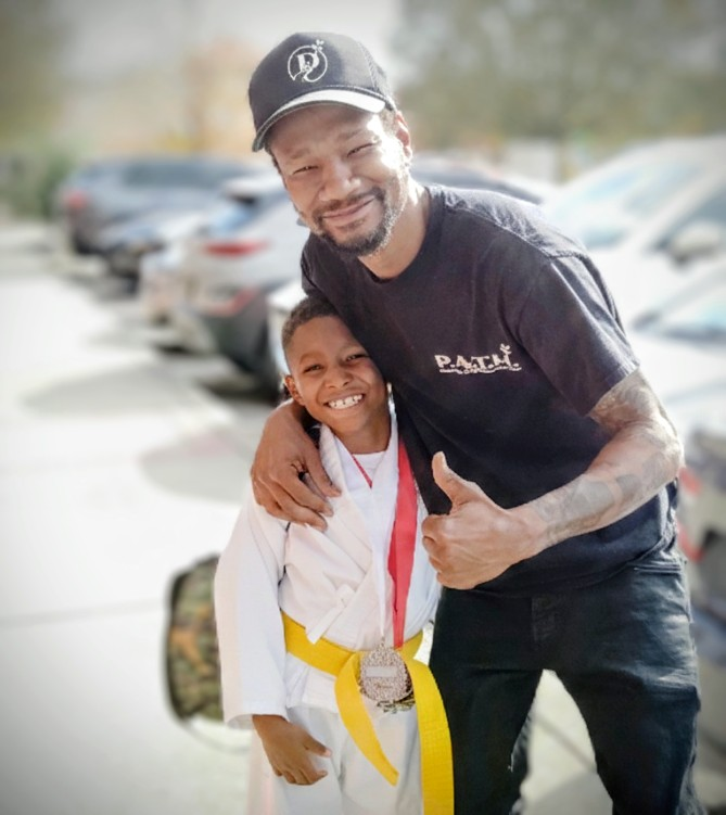Smiling coach hugs a child in a white martial arts uniform with a yellow belt outdoors.